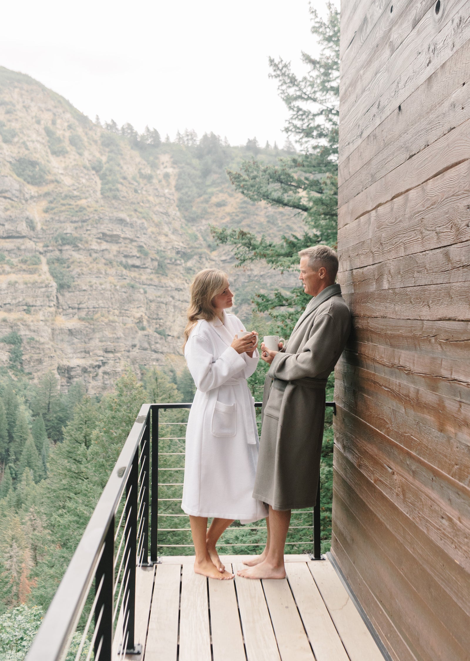 Man and woman standing on out door porch. In the background you can see a mountain and trees. The woman wears a white bathrobe and the man wheres the charcoal robe. The man and woman are holding mugs and talking with each other. 