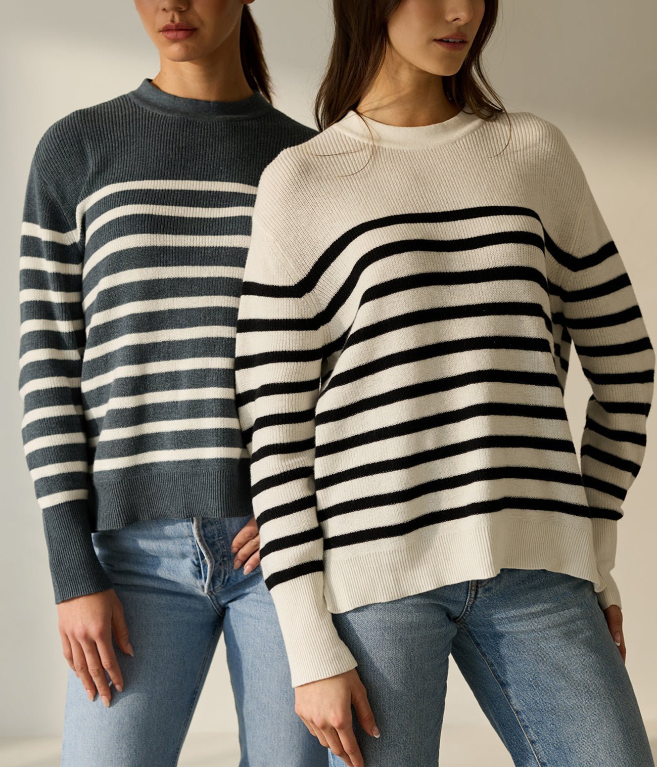 Two people wearing blue and white striped sweaters stand side by side. They have long hair and are dressed in light blue jeans. The background is neutral and softly lit.