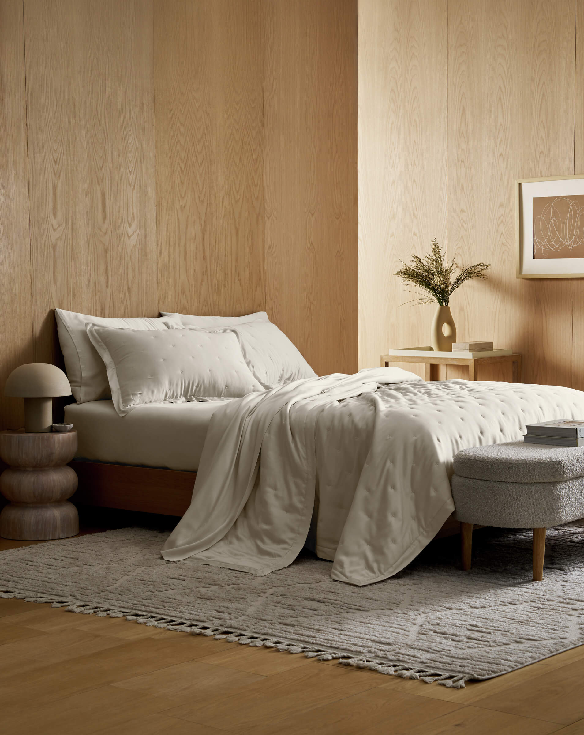 A cozy, modern bedroom with a wooden bed, cream bedding, a textured rug, a small nightstand with a vase, a round ottoman, and wooden walls decorated with framed art.