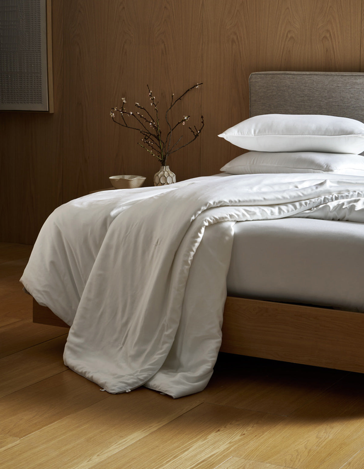 A neatly made bed with white bedding and pillows, a wooden headboard, and a nightstand with a vase holding branches, set against wood-paneled walls and a light wood floor.