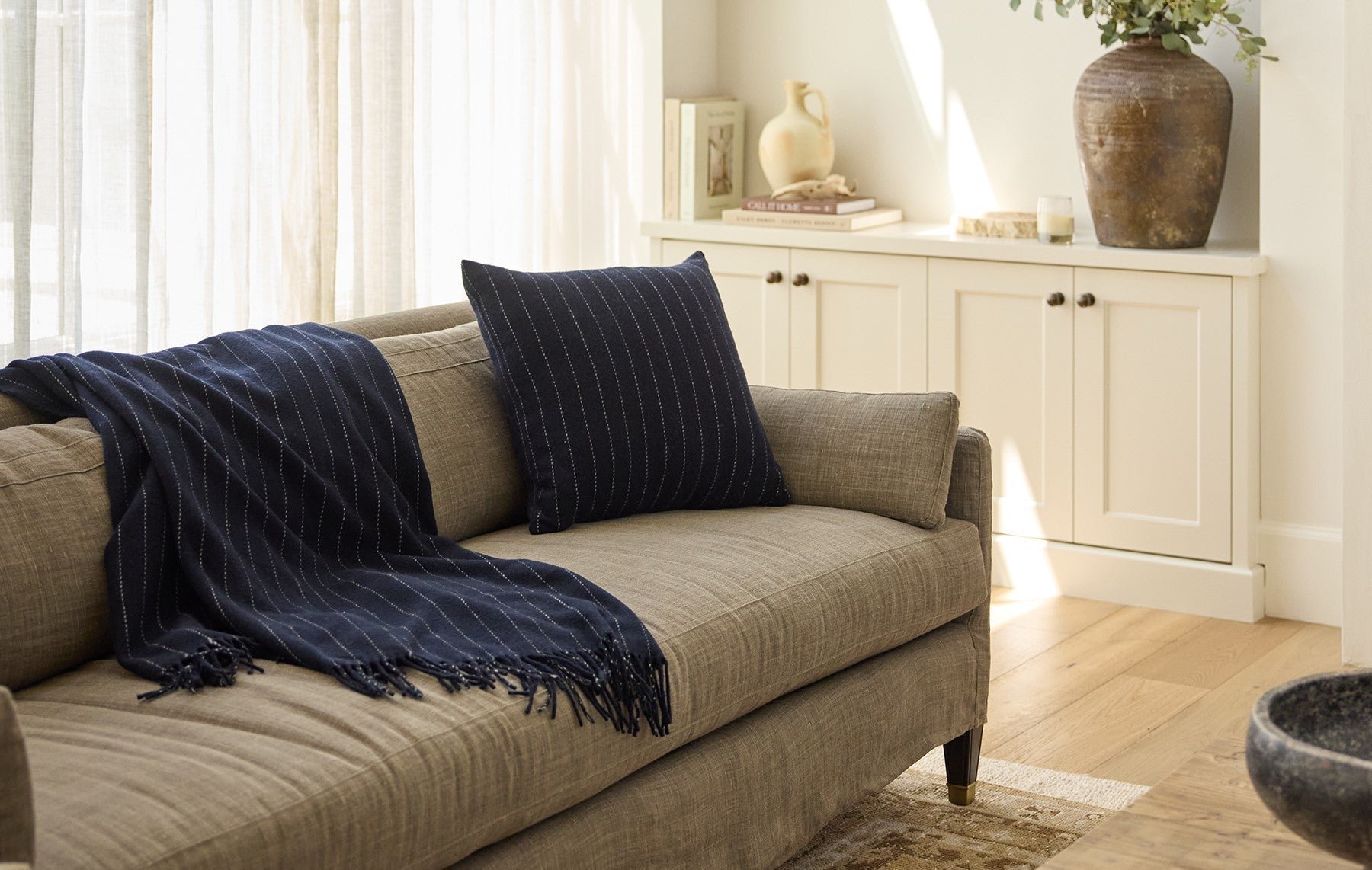 A beige sofa with a dark blue striped throw and matching pillow. Soft sunlight streams through sheer curtains, illuminating a bookshelf with books and decorative vases in the background.
