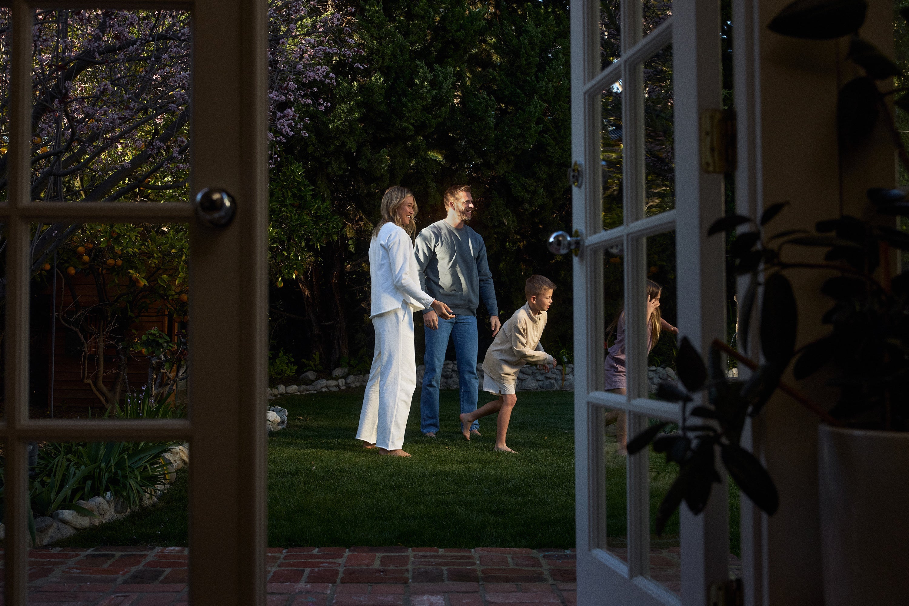 A family of four enjoys time together in a backyard garden, viewed through open glass doors. The parents smile while the children play on the grass surrounded by trees and plants.