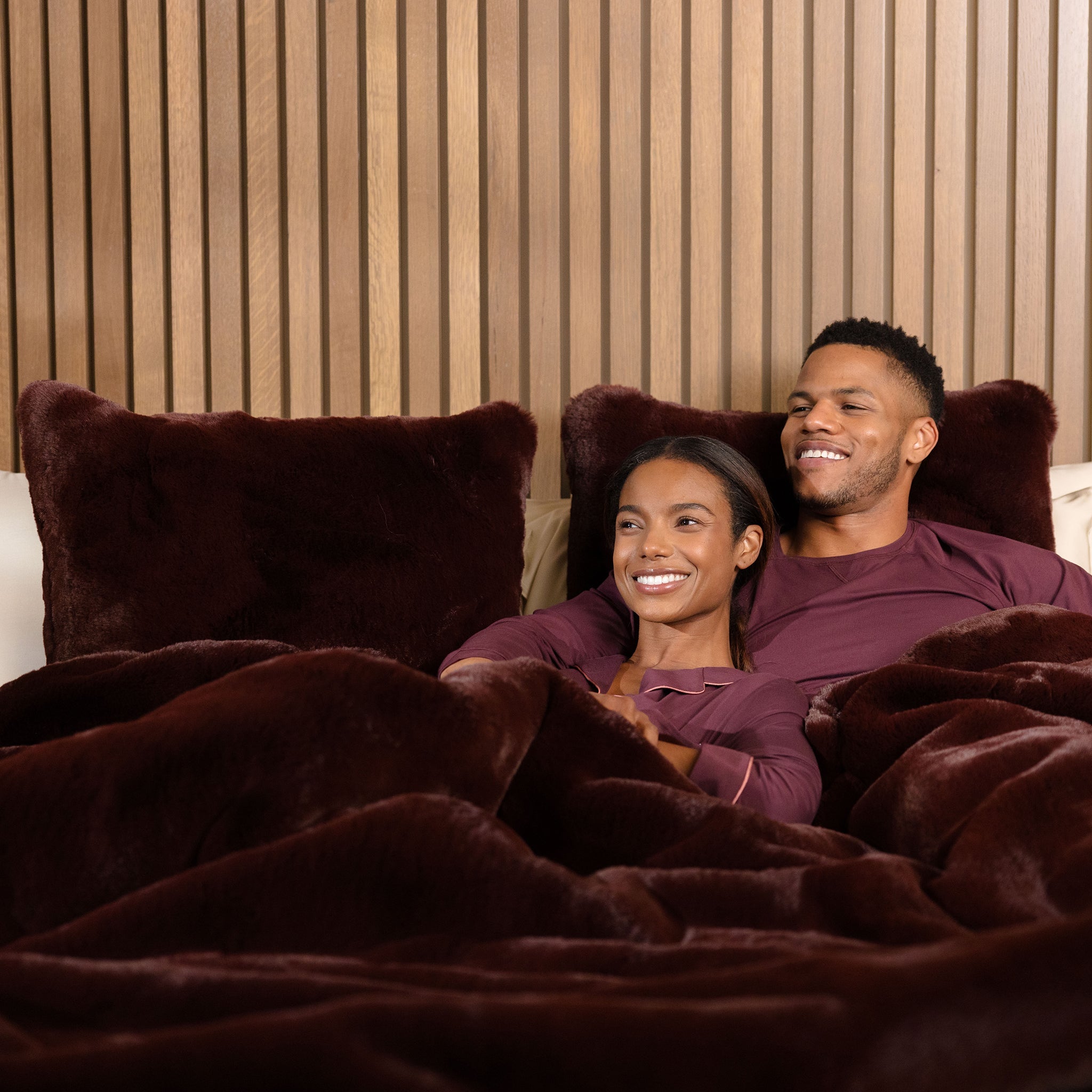 A smiling couple in matching maroon pajamas relaxes in bed under a dark, plush blanket. They are enjoying each other's company against a wooden, slatted backdrop.