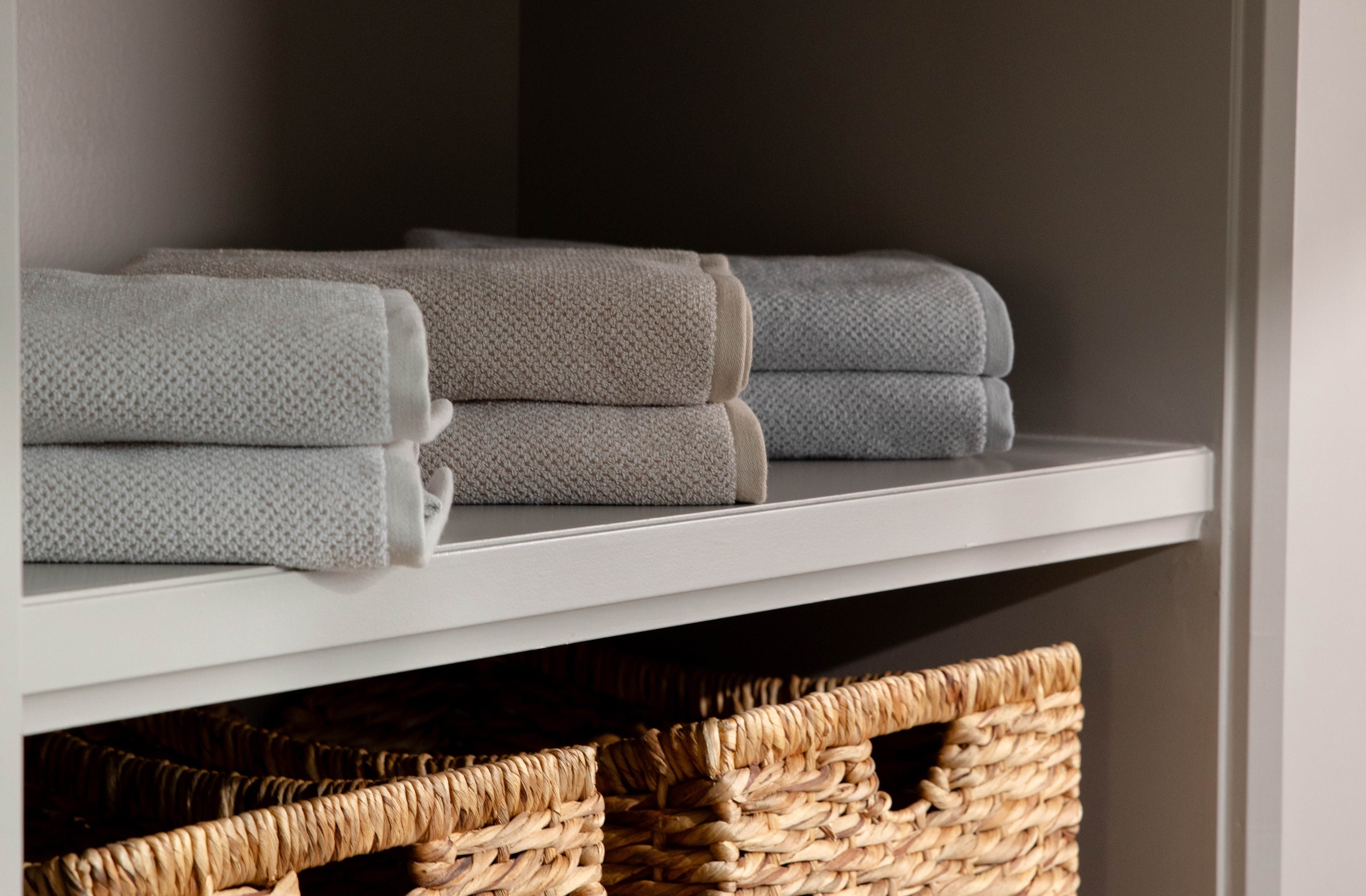 Folded towels in shades of gray and beige are neatly stacked on a white shelf. Below the shelf, two woven baskets are positioned on the next shelf down. The background is a neutral-colored closet or storage unit.