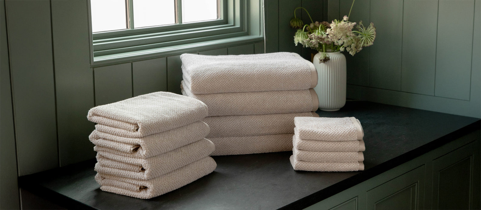 A stack of Cozy Earth Nantucket Bath Towels, Hand Towels, and Washcloths sit on a countertop under a window. A vase with flowers and green paneled walls are also shown.