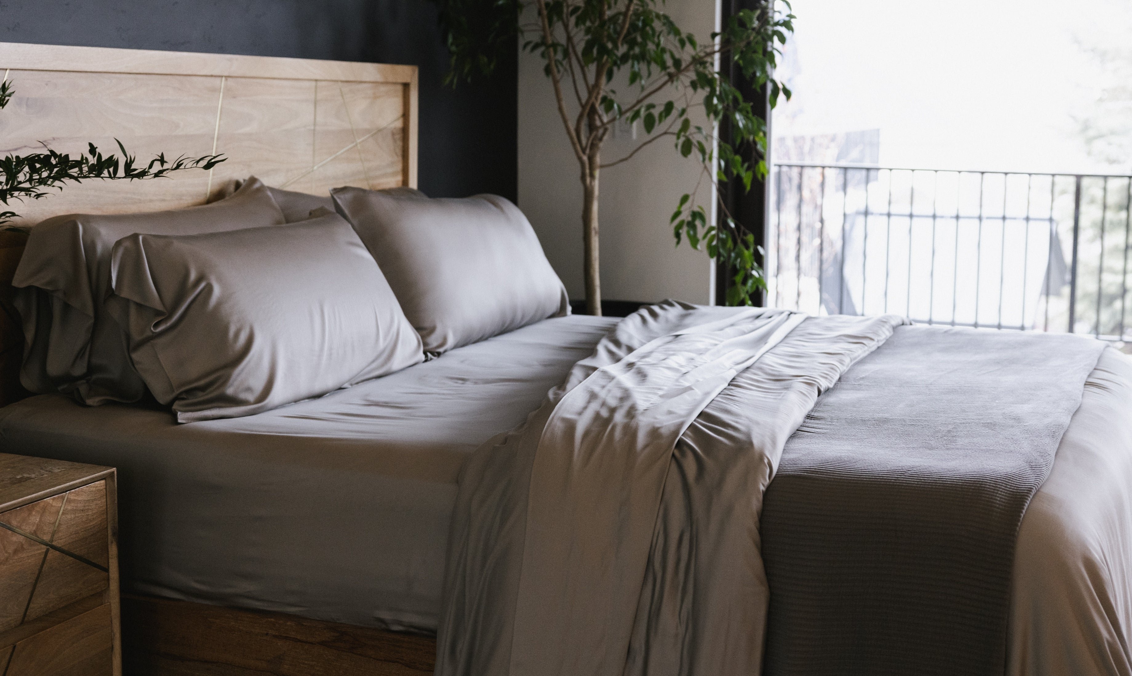 A neatly made bed with grey sheets and several pillows is situated in a modern bedroom with wooden furniture. A large window lets in natural light, and there is a tall plant next to the bed.