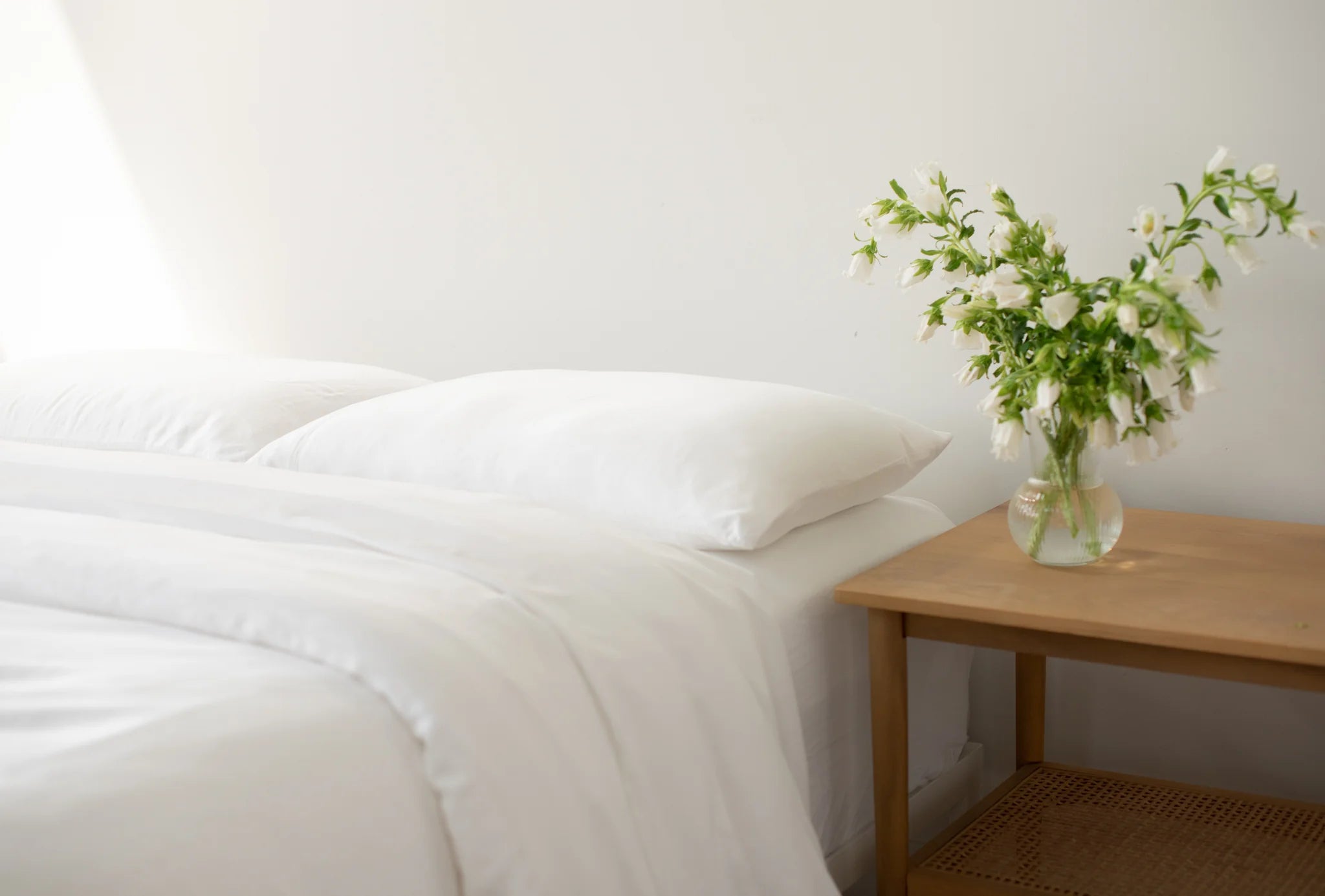 A neatly made bed with white linens and pillows is beside a wooden nightstand holding a clear vase filled with white flowers. The background is a plain white wall.