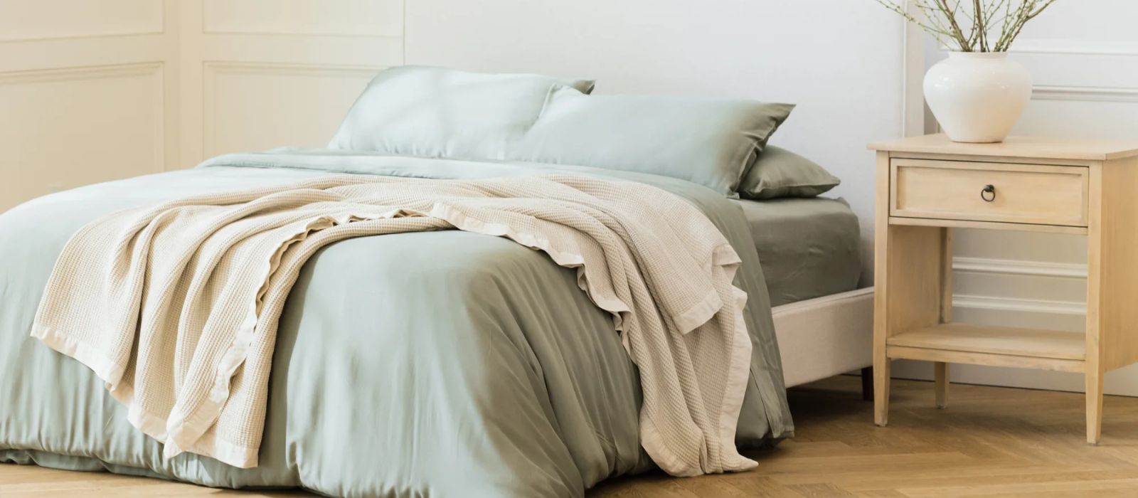 A minimalist bedroom features a bed with soft green bedding and a beige throw, next to a light wood nightstand holding a white vase with branches. The room has light-colored walls and a wooden floor.