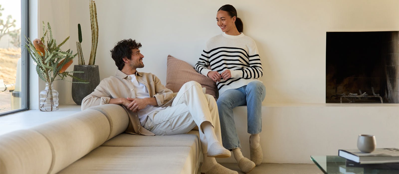 A man and woman sit in a living room wearing Cozy Earth apparel. A cactus and greenery are on a shelf behind them. A window lets in light.