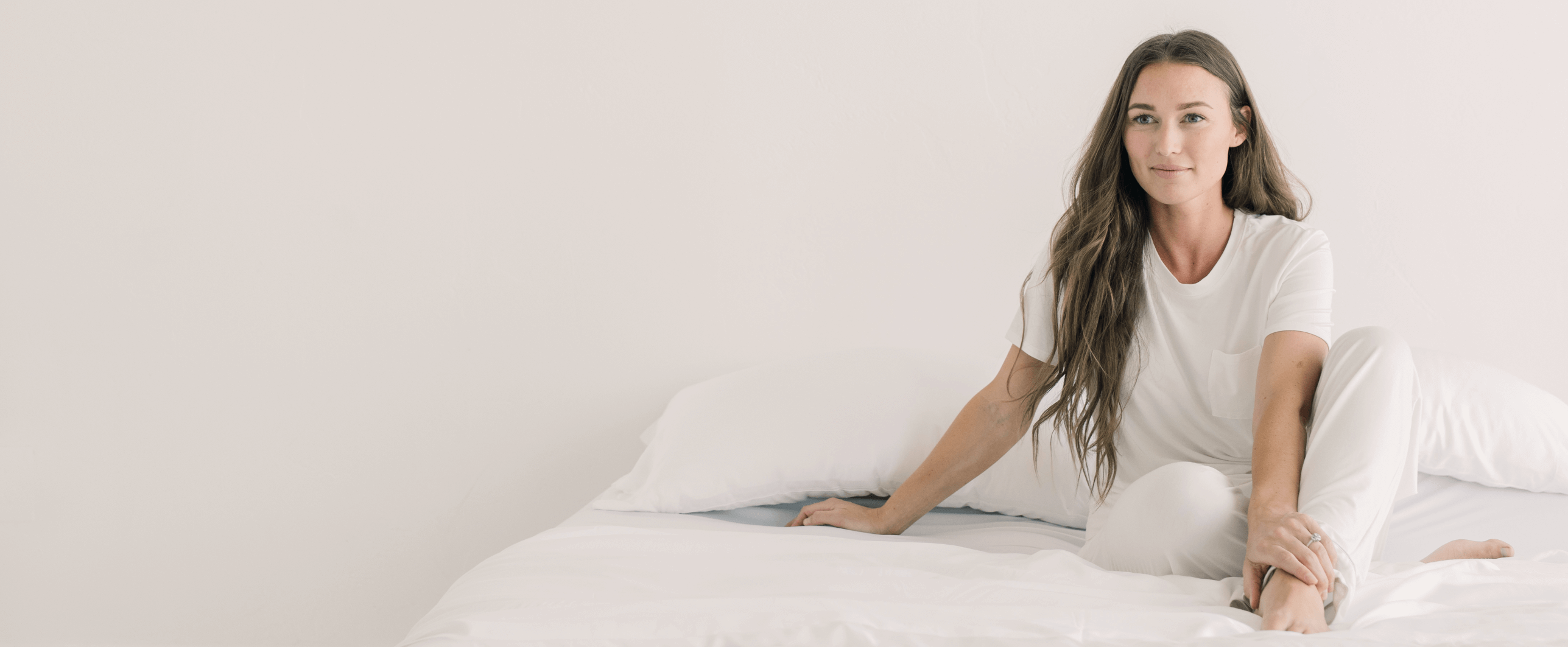 A woman with long hair sits on a bed in a bright, minimalist room. She wears a white outfit and leans slightly against a pillow, gazing towards the camera.