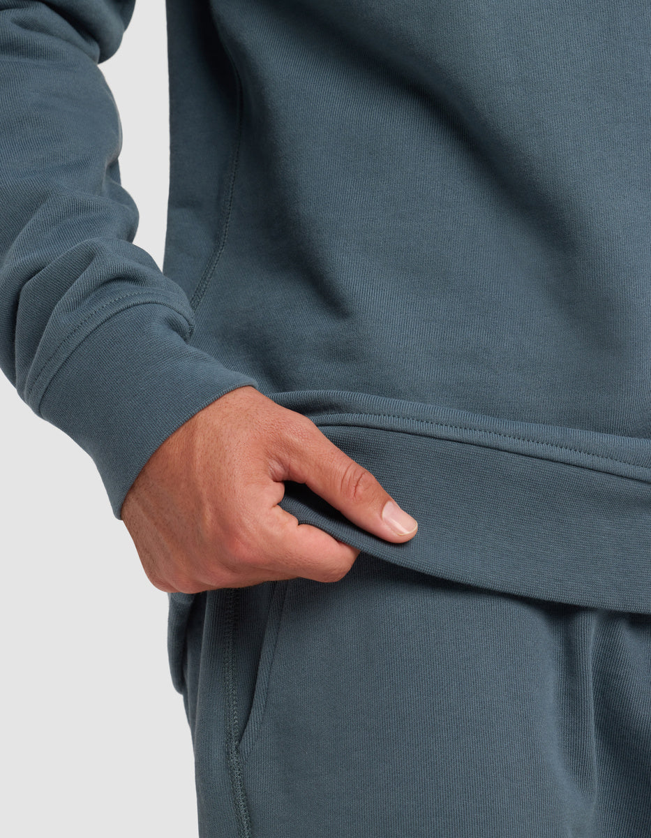 A person wearing a blue-green sweatshirt and matching sweatpants holds the bottom hem of the sweatshirt with their right hand, showing the texture and fit of the clothing.