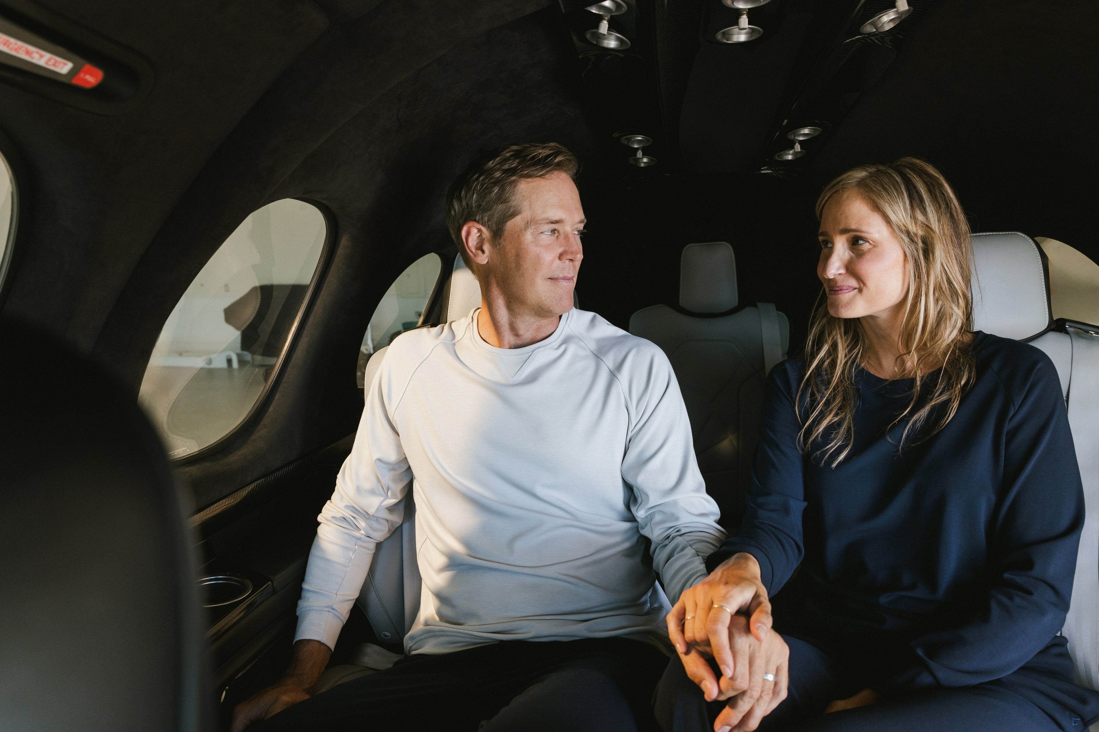 A couple sits together inside a private jet, holding hands and looking at each other with soft smiles, dressed in casual light-colored clothing.