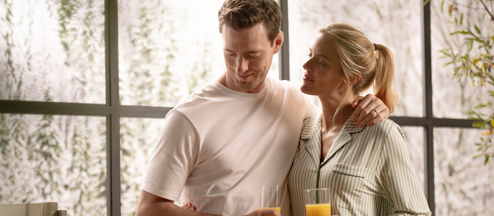 A woman looks at a man who has his arm around her. They stand in front of a window overlooking a winter scene