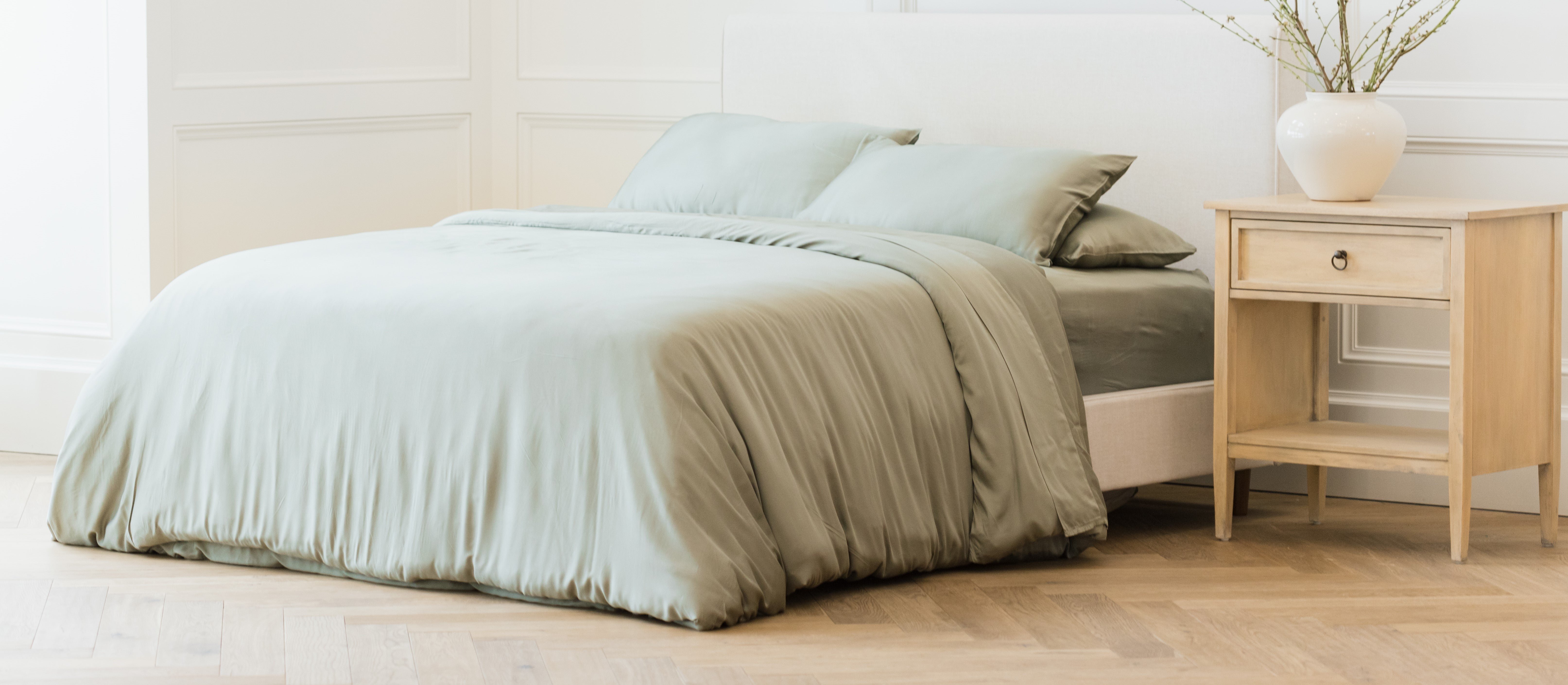 A neatly made bed with light green bedding in a bright, minimalistic bedroom. Next to the bed is a wooden nightstand holding a white vase with branches. The room has light-colored walls and wood flooring.
