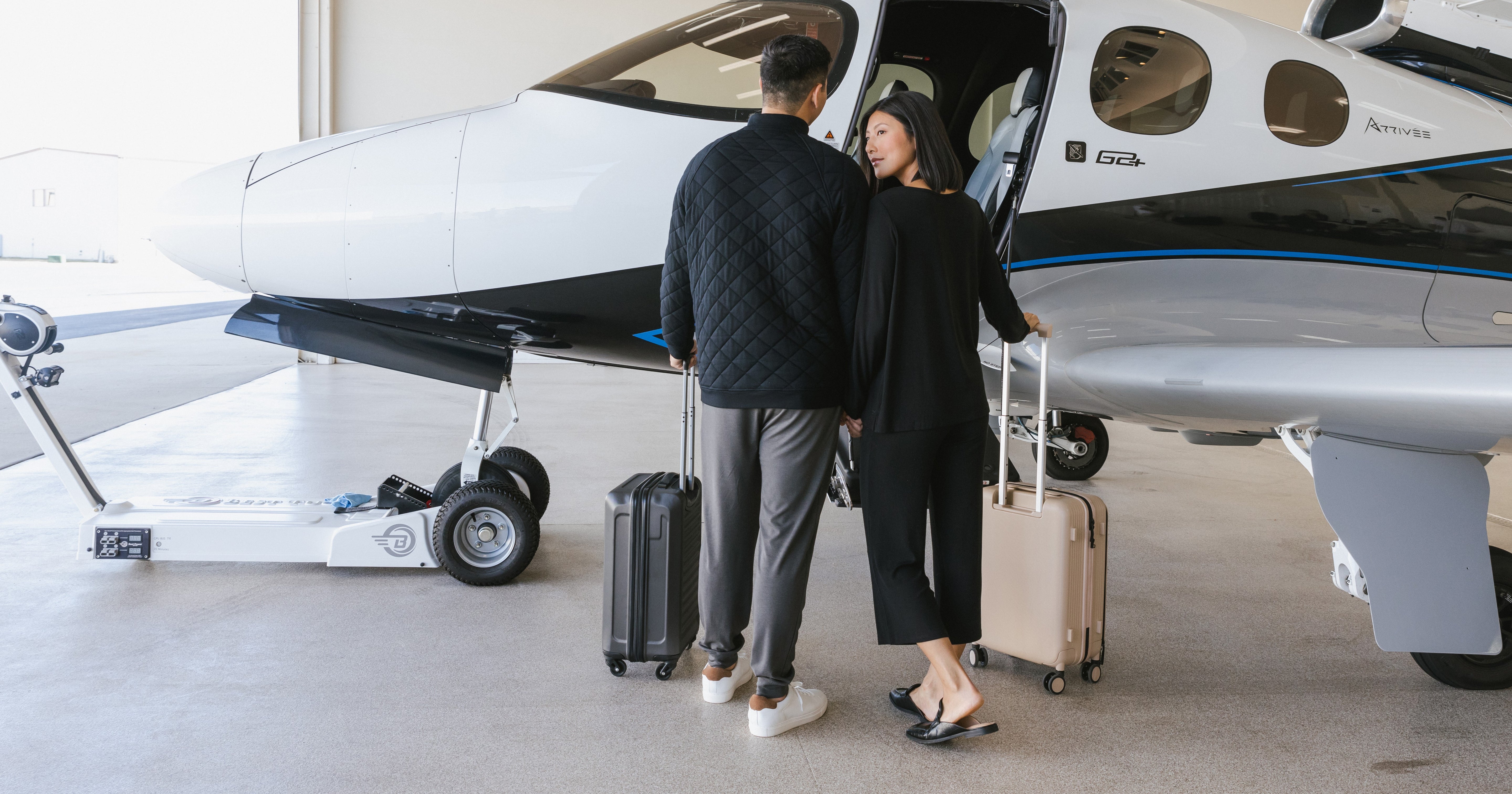 A man and woman, each with a suitcase, stand near the open door of a small private jet, preparing to board. They are casually dressed and appear to be traveling together.