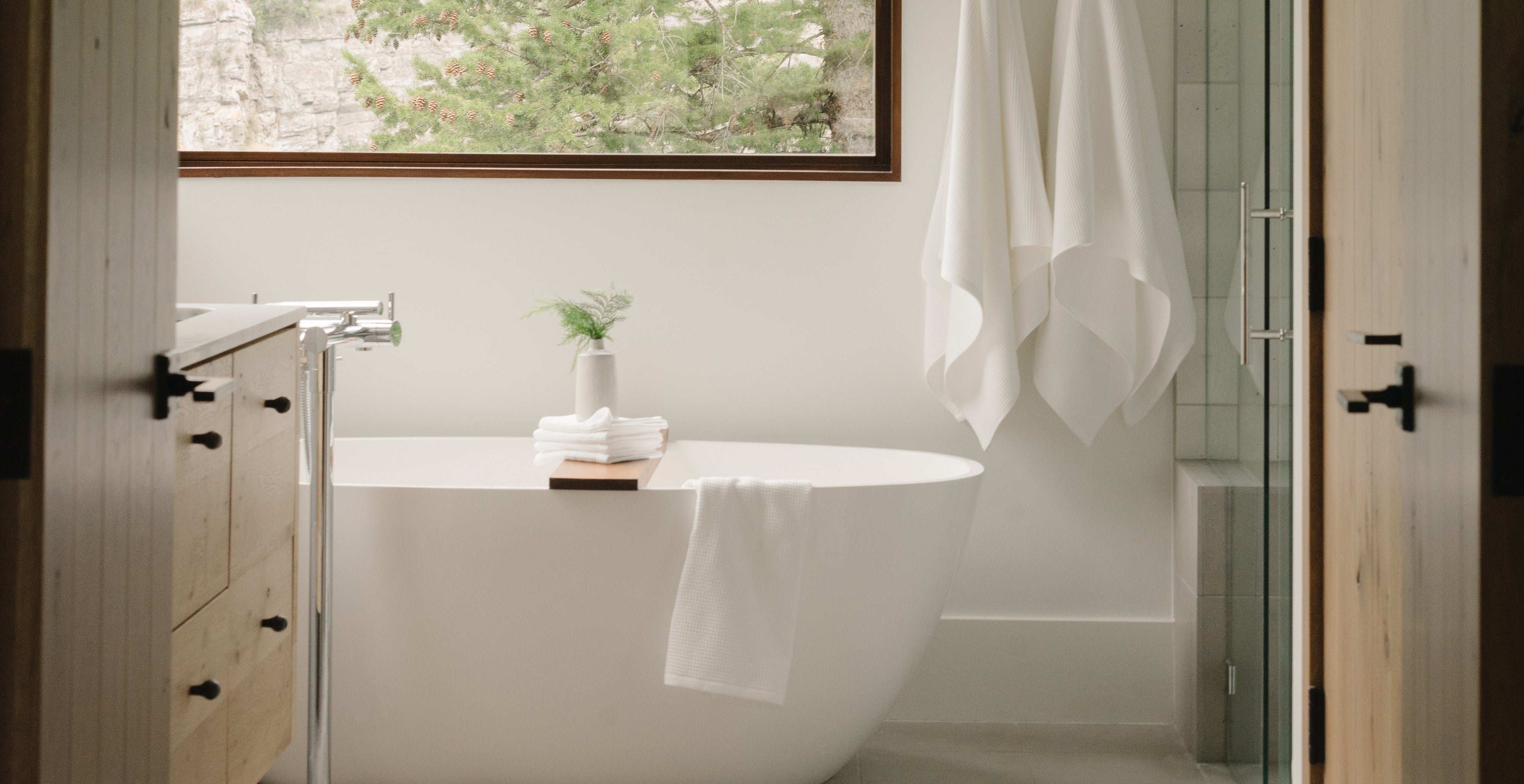 Minimalist bathroom with a freestanding white bathtub, a small plant on a wooden bath tray, and towels. Large window with a view of trees and natural light enhances the serene atmosphere.