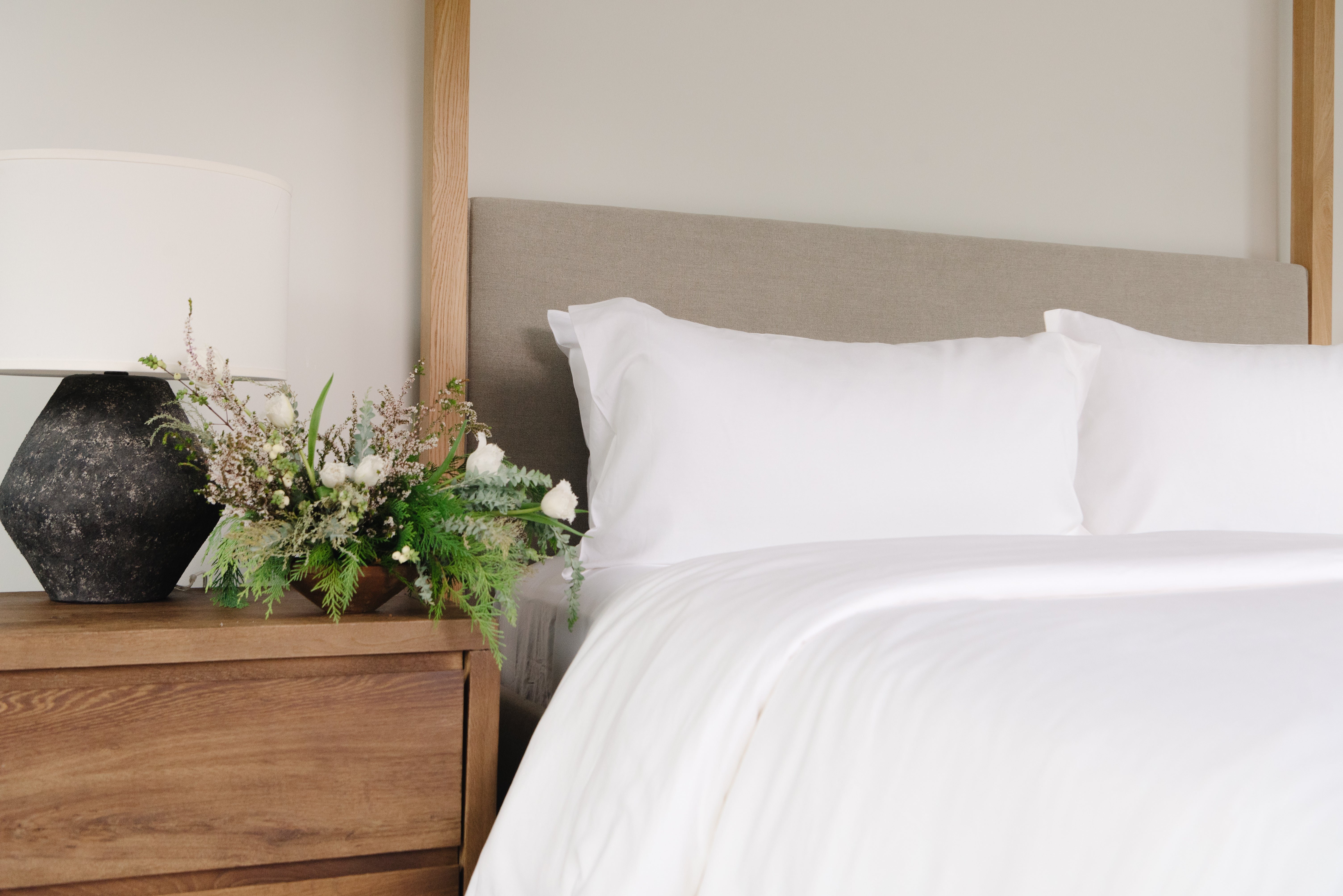 A neatly made bed with white linens is next to a wooden nightstand. The nightstand holds a black geometric lamp and a floral arrangement with green and white flowers. The headboard is upholstered in light gray fabric.