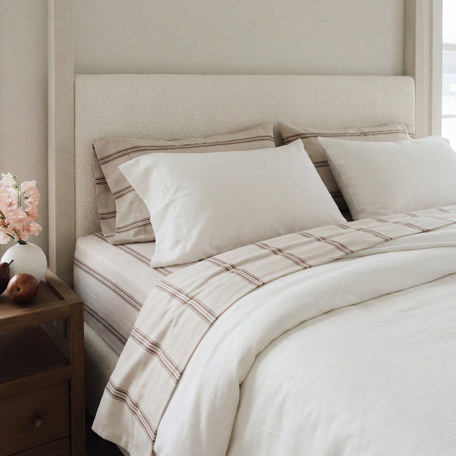 A neatly made bed with beige and white striped bedding, two large pillows, and a light blanket. A wooden nightstand beside the bed holds a small vase of pink flowers and some fruit.