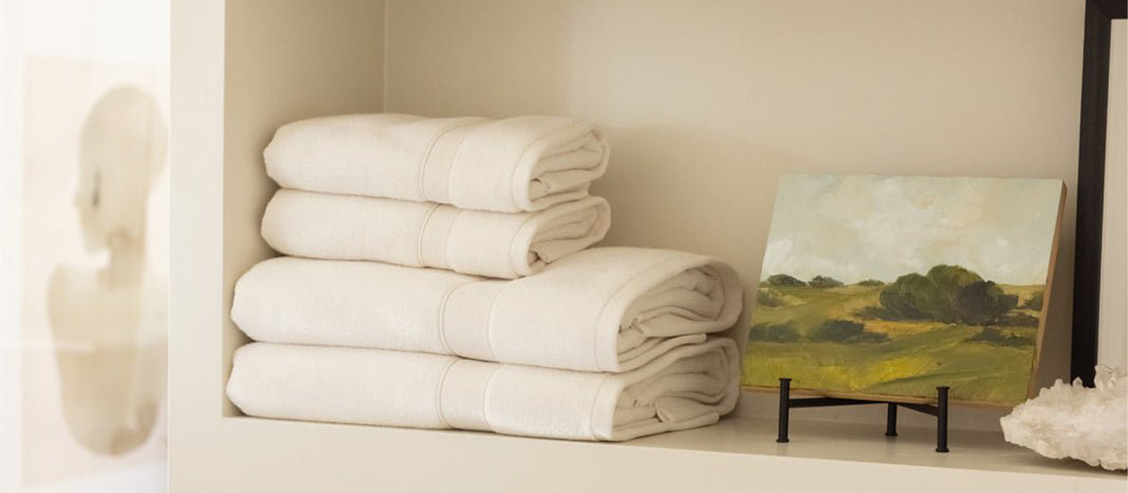Three neatly folded white towels sit on a shelf next to a small landscape painting on a stand, with soft, neutral lighting and a blurred artwork in the background.
