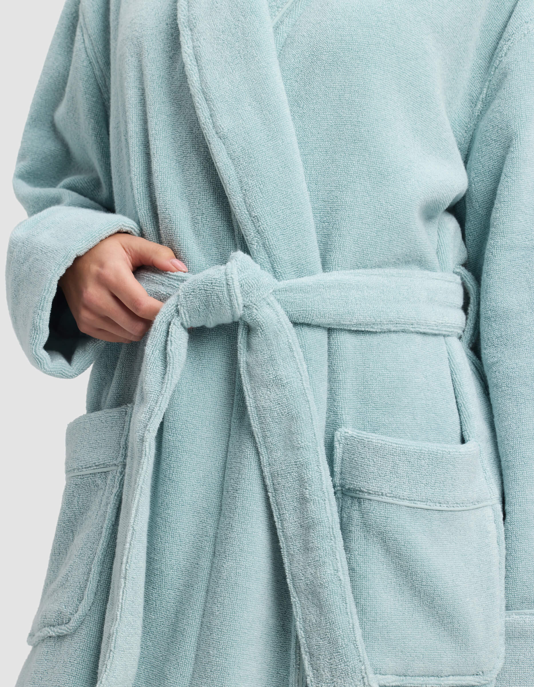 A person wearing a light blue bathrobe ties the belt at the waist. Only the torso and hands are visible, with two large front pockets on the robe.