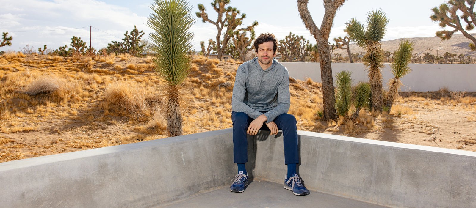 A man sits on a low concrete wall in a desert landscape.