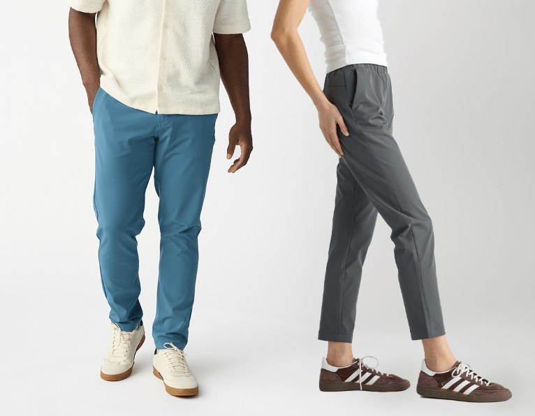 A man in a cream shirt and blue pants smiles, standing beside a woman in a white top and gray pants. Both wear sneakers and pose against a plain white background.