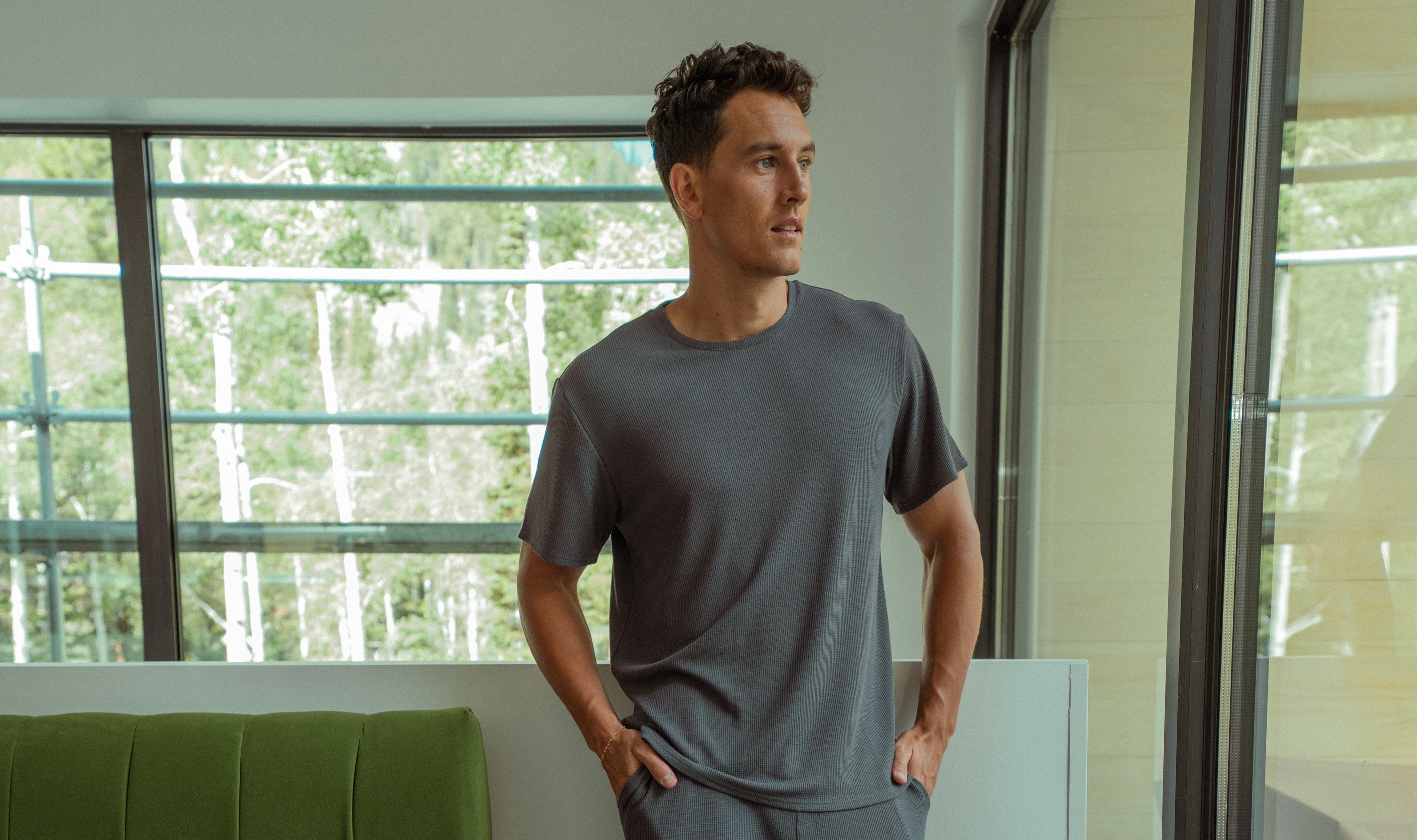 A person wearing a gray t-shirt stands indoors by a large window. They have their hands in their pockets and look to the side. The background features green trees visible through the window.