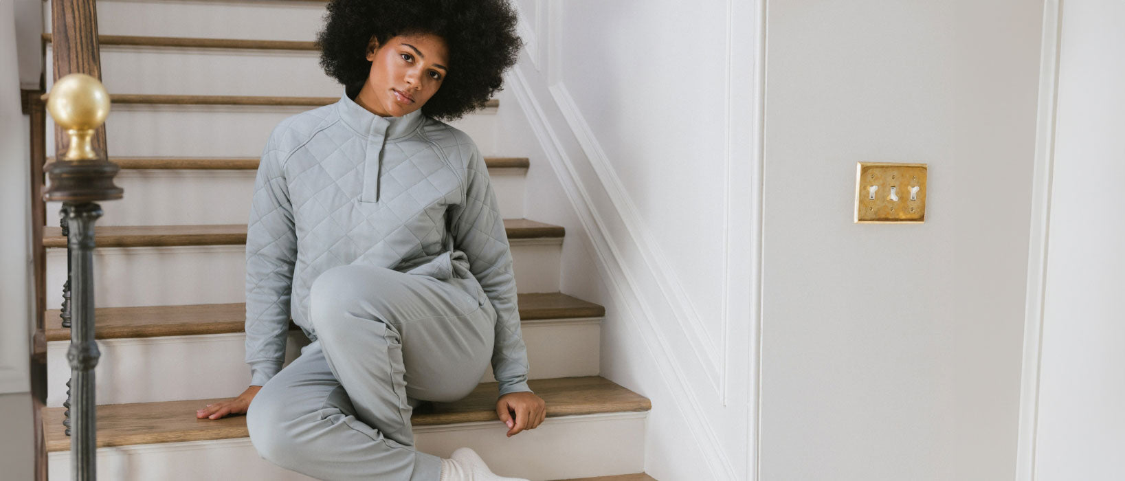 A person with curly hair and a neutral expression sits on indoor wooden stairs, wearing a light gray quilted top and matching pants, with white socks.