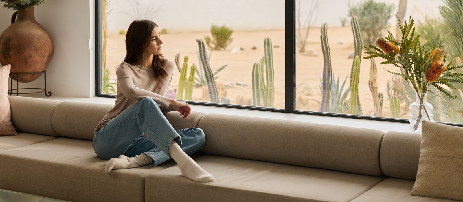 A woman in jeans and a tan shirt sits on a couch and looks out a window at a desert landscape