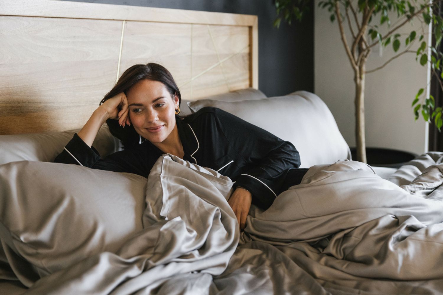 A woman in black pajamas lies on a bed with grey sheets, propped up on one arm, smiling softly. A wooden headboard and a leafy indoor plant are visible in the background.