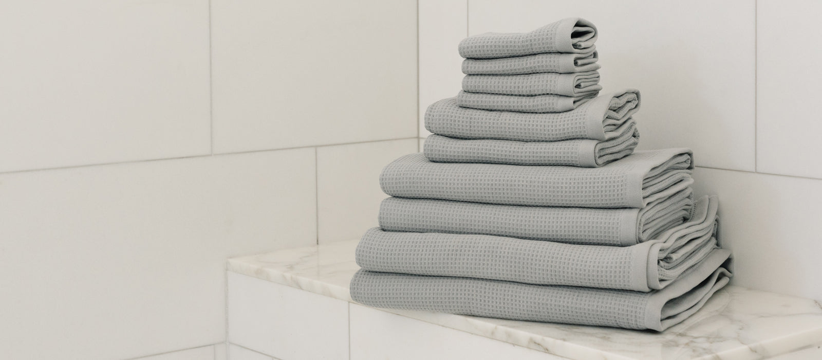 A stack of light blue Cozy Earth Waffle Bath Towels sit on a tiled bench in a shower