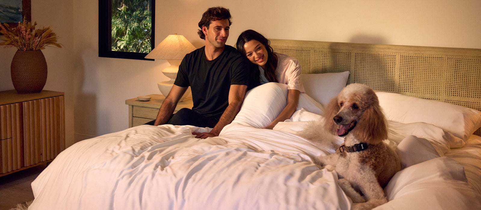 A man and woman sit in bed with a poodle lounging at the foot of the