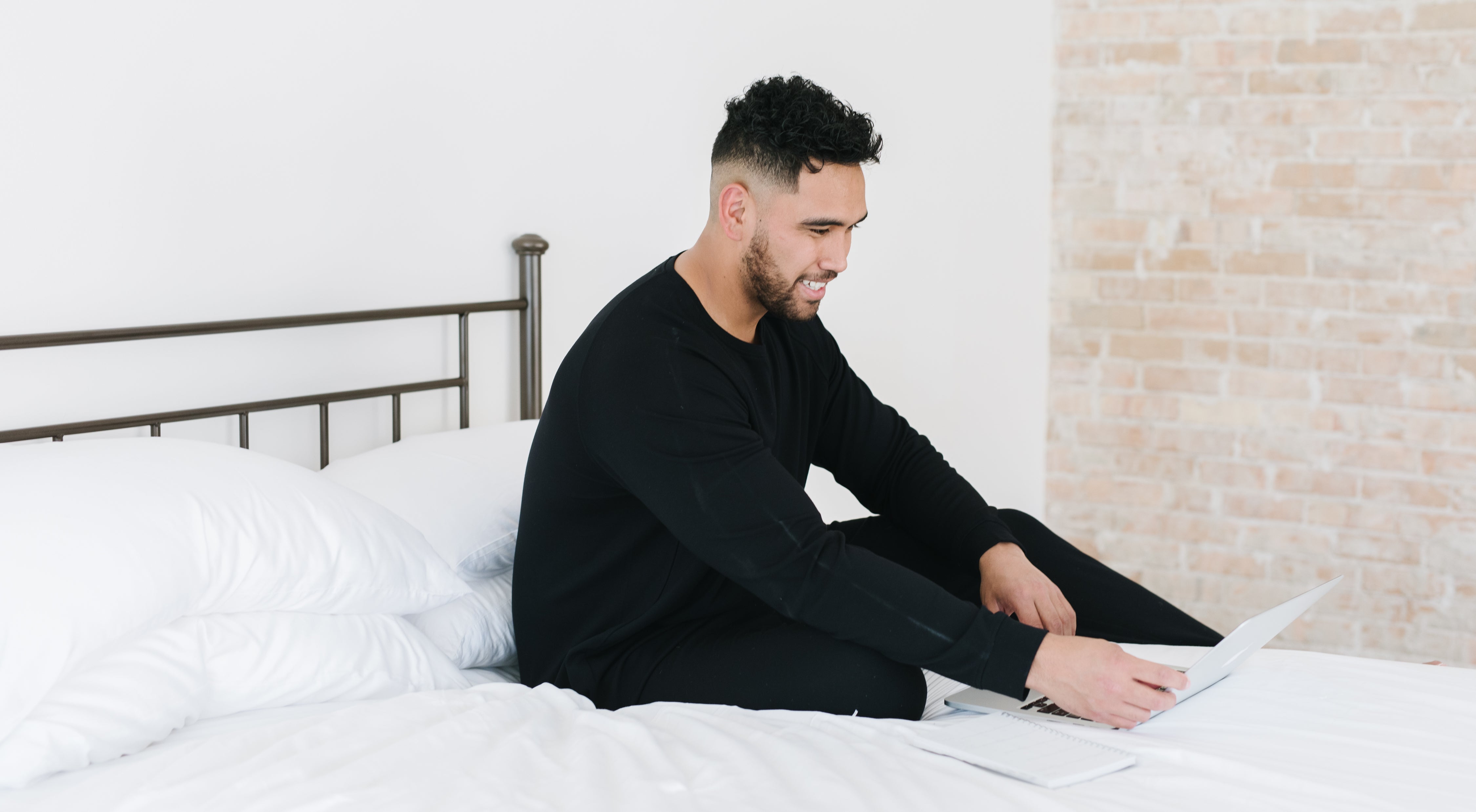 A man dressed in black sits cross-legged on a white bed, using a laptop. The room has a minimalist style with a brick wall and a metal bed frame. He appears focused on the screen.