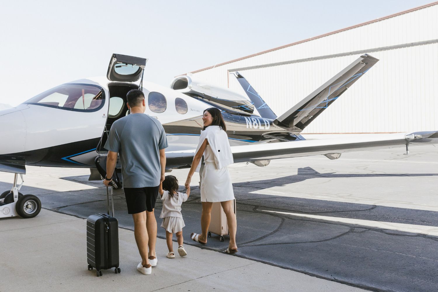 A family of three, with a man carrying luggage, a woman in a white dress, and a small child, walks toward a private jet on an airport tarmac.