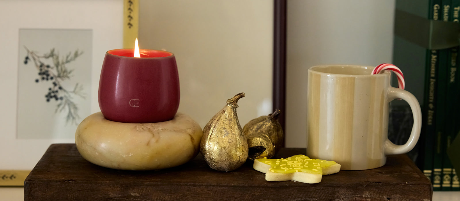 A lit candle sits on a shelf next to a mug, golden pear, and sugar cookie