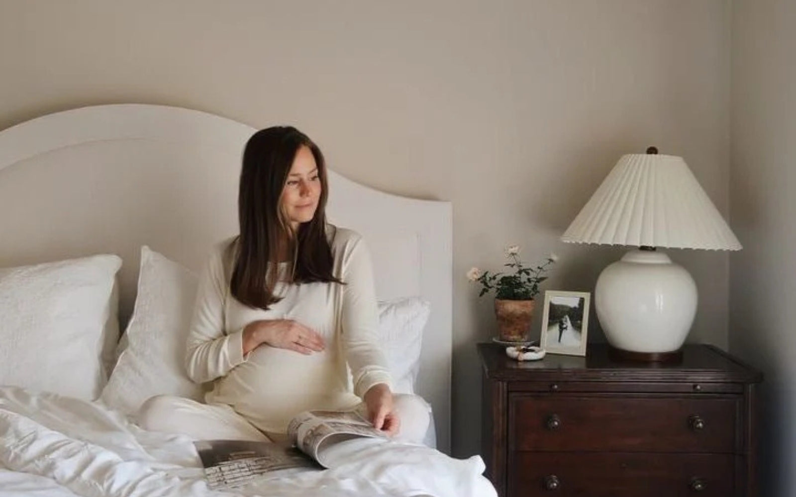 A pregnant woman wearing cream pajama set sits in bed with her hand on her tummy