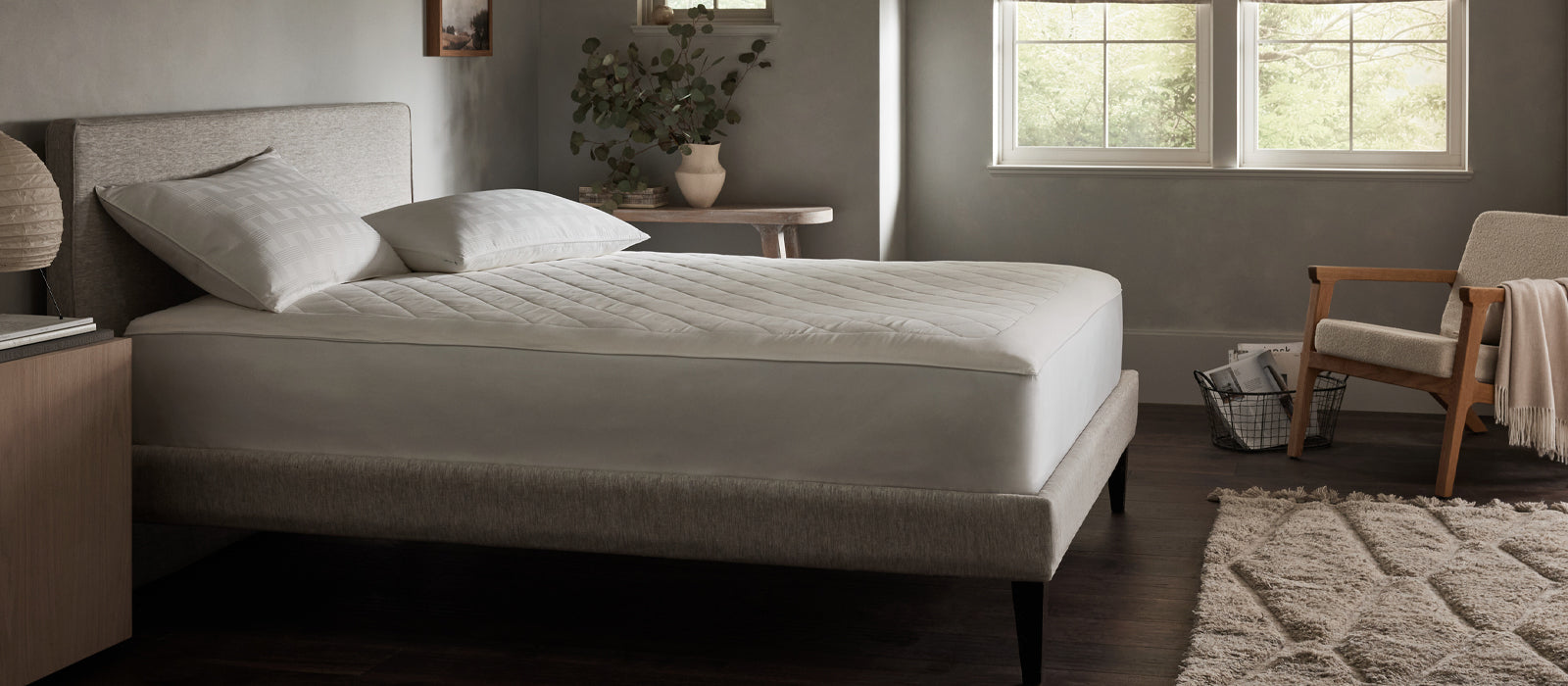 A neatly made bed with white bedding sits in a softly lit, modern bedroom with a rug, wooden chair, and a window letting in natural light.