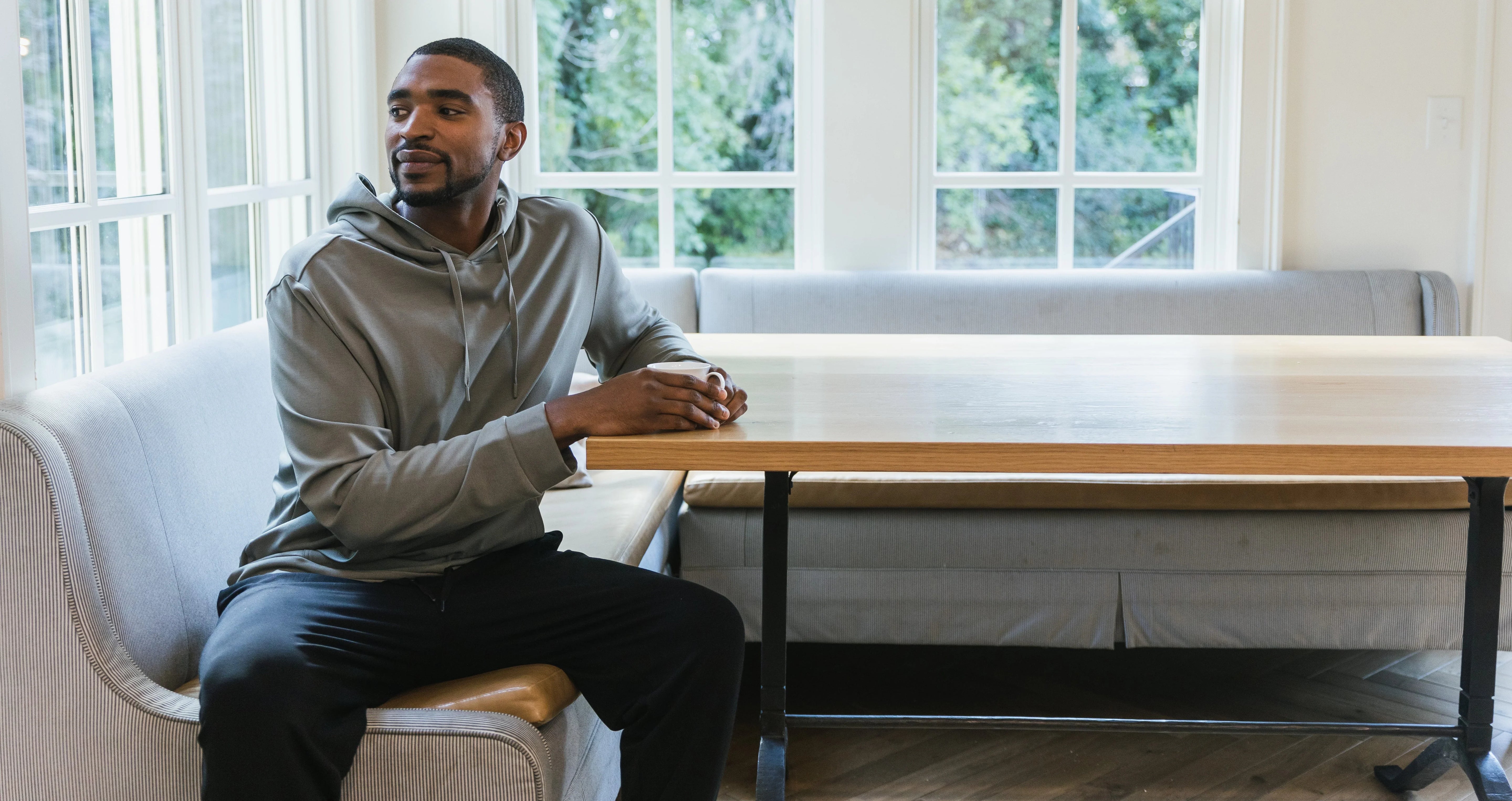 A man in a gray hoodie sits on a light gray bench at a wooden table, looking to his left. Large windows behind him reveal greenery outside.