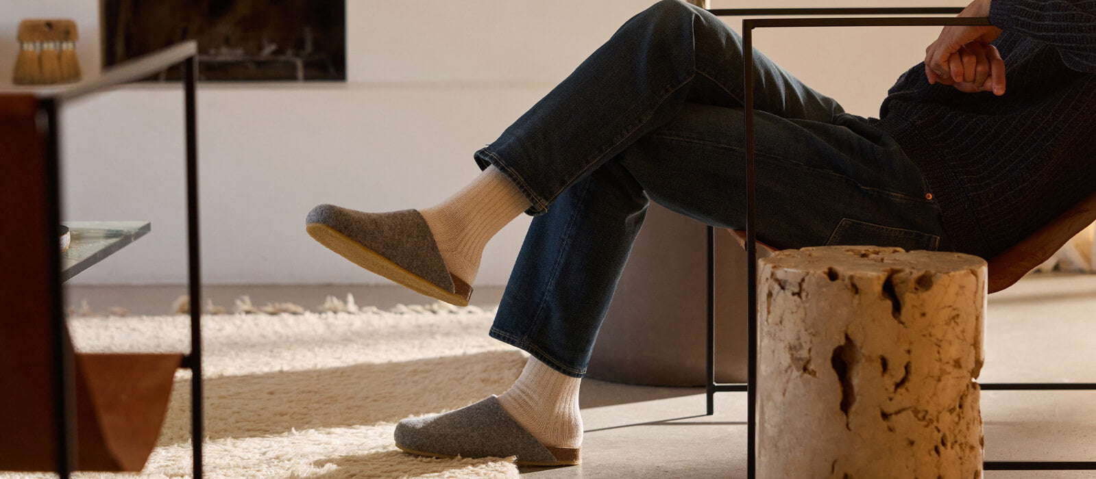 A person wearing jeans, a dark sweater, white socks, and grey slippers sits sideways in a modern chair beside a rustic wooden stump in a cozy, sunlit room.