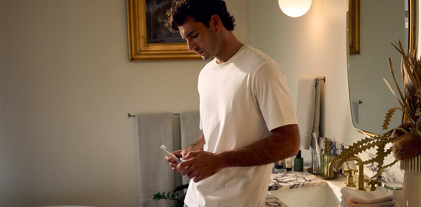 A man wearing a white tee stands in a bathroom holding a toothbrush