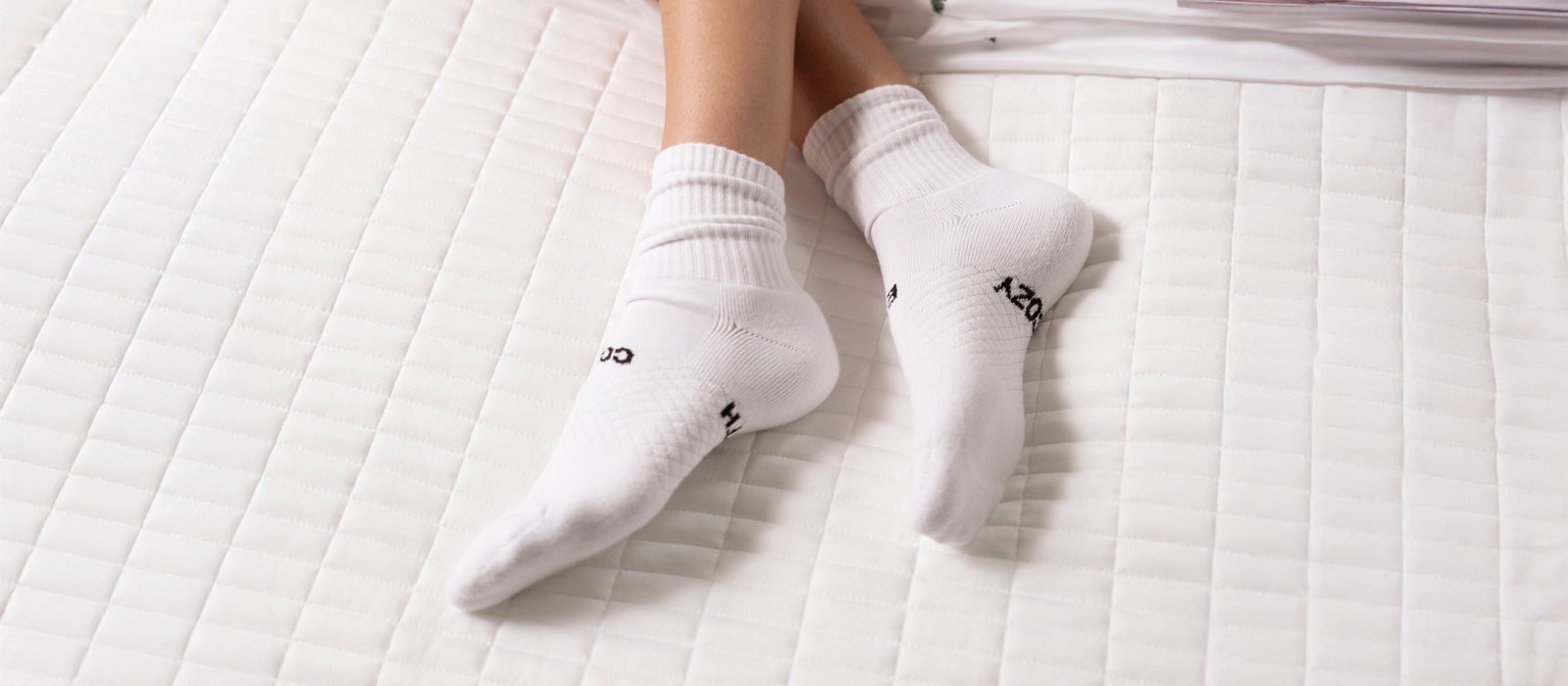 A person wearing white socks lies on a white, quilted bedspread with their feet crossed at the ankles. The socks have black text near the toes.