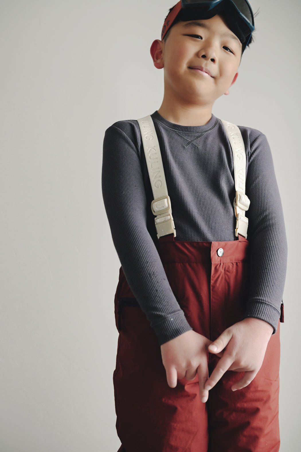 A young boy smiles with hands clasped, wearing the Cozy Earth Children's Bamboo Waffle Knit Base Layer Long Sleeve and red snow pants, accessorized with a red headband, ski goggles, white suspenders, against a plain background.