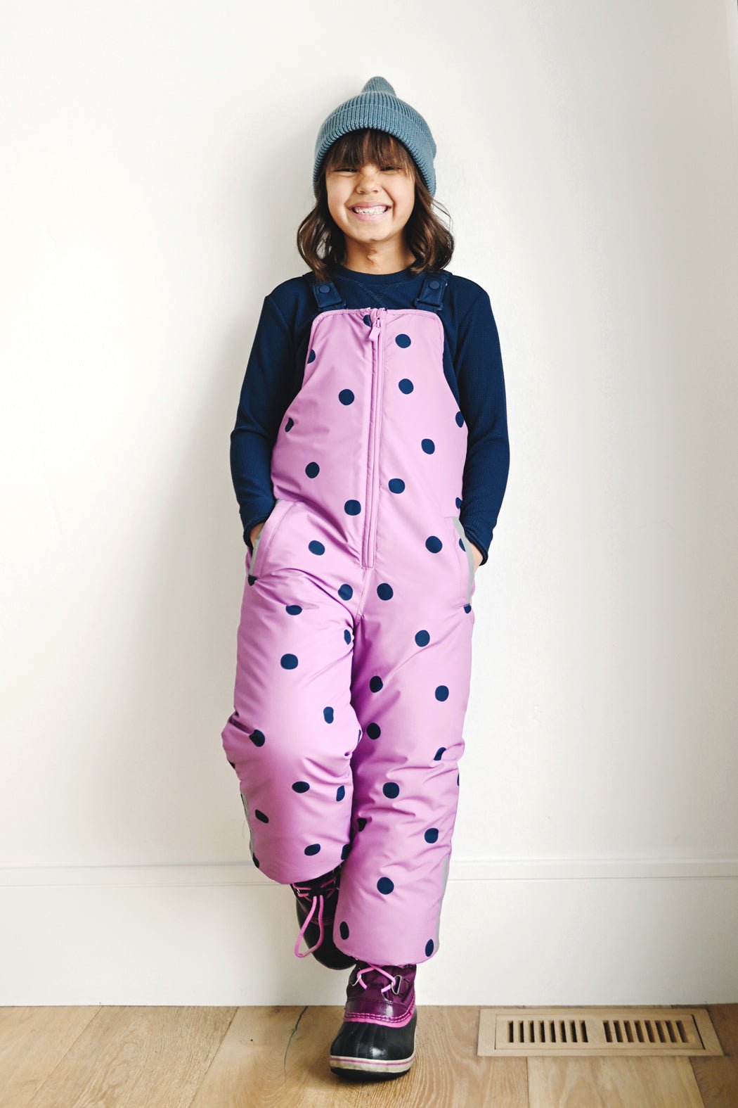 A smiling child in a Cozy Earth Children's Bamboo Waffle Knit Base Layer Long Sleeve and pink polka-dotted snow pants stands indoors, hands in pockets and one foot crossed over the other.
