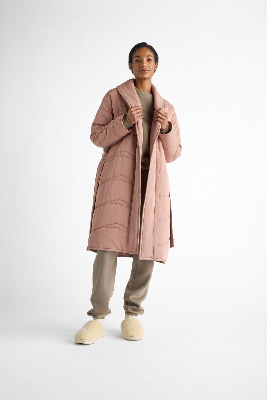 A person wears a Cozy Earth Women's Quilted House Coat in light pink over a beige outfit with tan fuzzy slippers, set against a plain white background. |Color:Rosewood