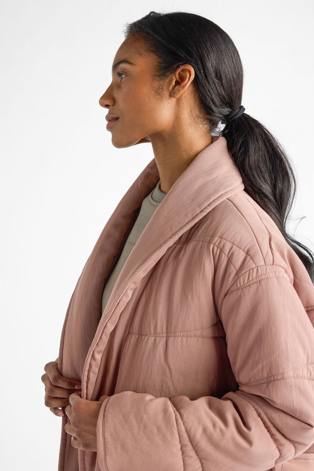 A person with long hair tied back, wearing Cozy Earth's Women's Quilted House Coat in light pink over a beige top, gazes to the left against a plain white background. |Color:Rosewood