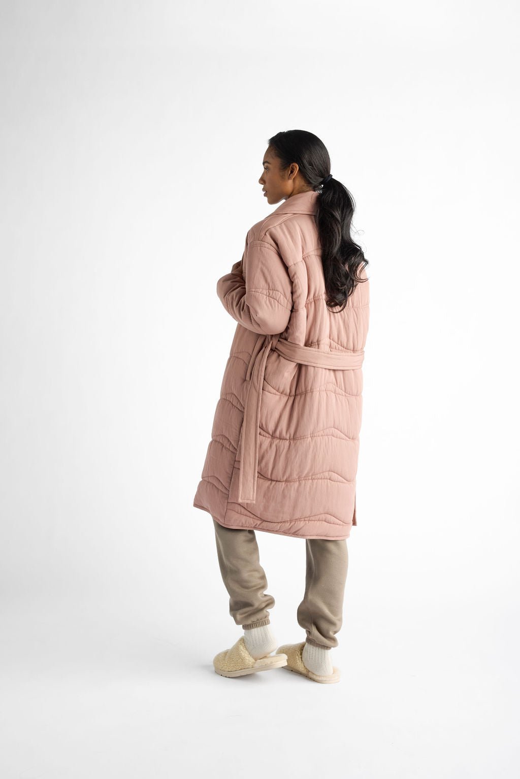 A person with long dark hair is in profile, wearing a pink Women's Quilted House Coat by Cozy Earth, beige pants, and light slippers against a plain white background. |Color:Rosewood