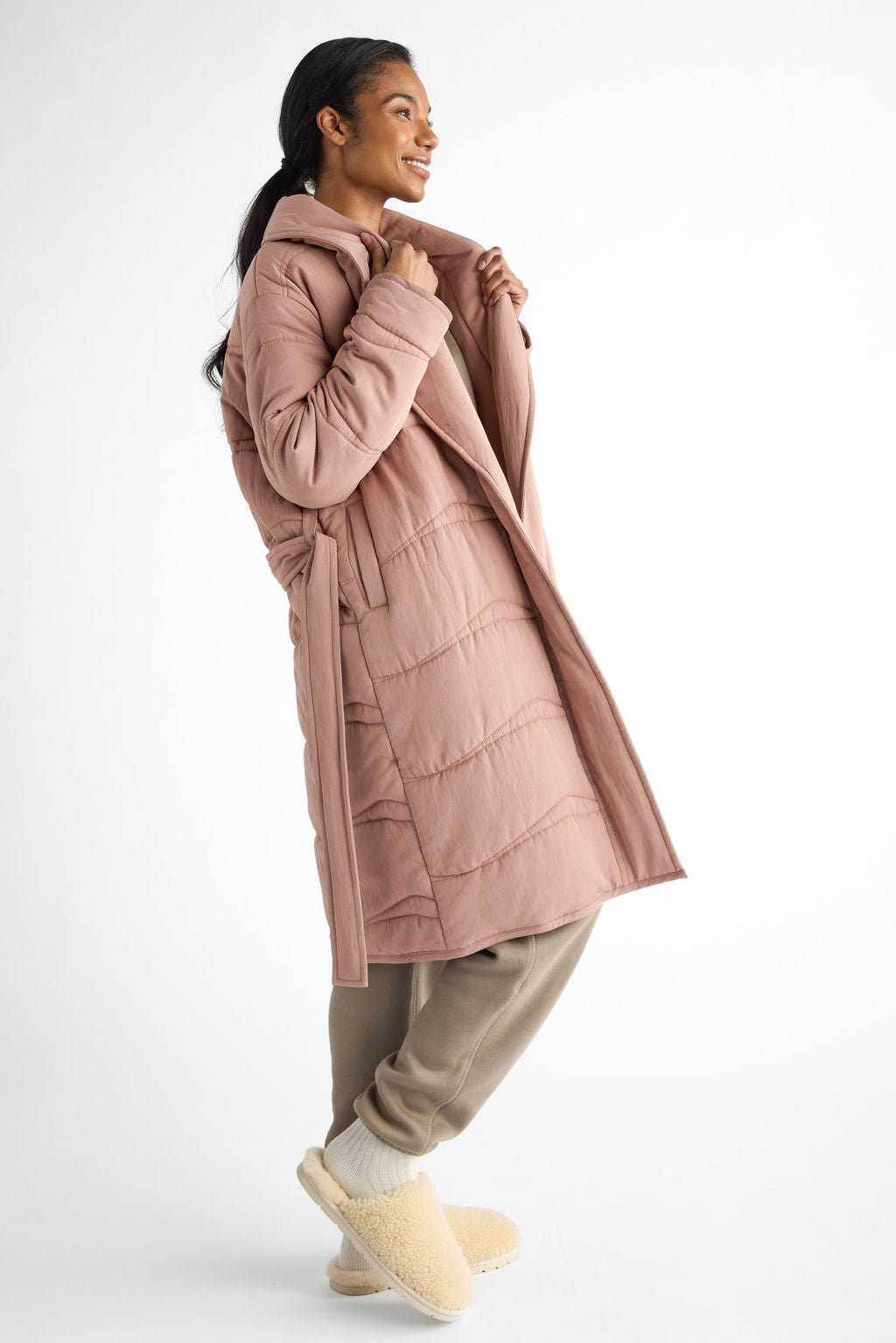 A person wearing a Women's Quilted House Coat by Cozy Earth in pink, paired with beige pants and fluffy beige slippers, smiles while holding the coat's lapel. They stand relaxed against a plain white background. |Color:Rosewood