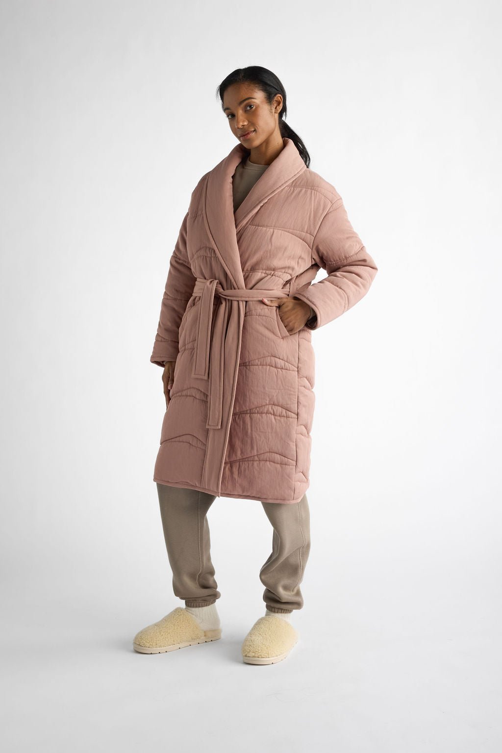 A person stands confidently against a plain white background, wearing Cozy Earth's Women's Quilted House Coat in long, pink with a tied belt, paired with beige pants and fluffy beige slippers. 