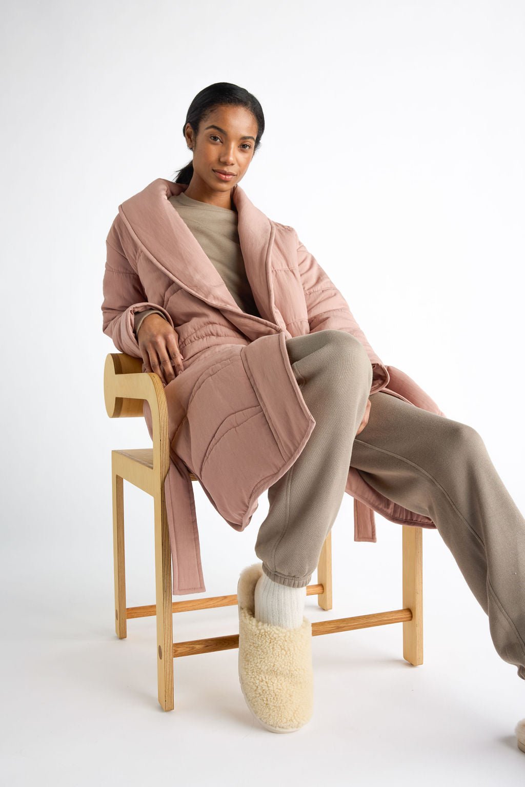 A person sits on a wooden chair in Cozy Earth's Women's Quilted House Coat, a beige sweater, and matching pants. With one leg crossed over the other, they wear cream-colored slippers against a plain white background. 