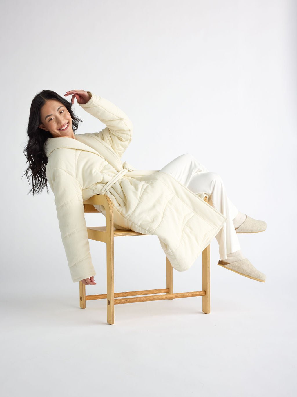 A woman in a Cozy Earth Women's Quilted House Coat and matching slippers smiles while lounging sideways on a wooden chair against a plain white background, highlighting the relaxed, comfortable atmosphere. 